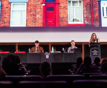 Three speakers on a stage, one standing behind a lectern, the others behind a table. A brick house with a red door is displayed on a screen behind them.