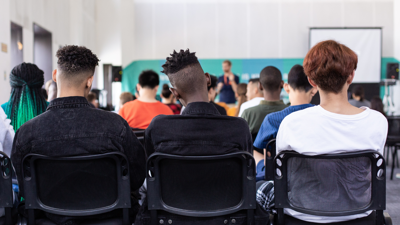 several rows of students sit as they listen to their teacher present. The back row which holds three students sits in the foreground.