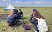 Five people dressed in outdoor gear and jackets squatting in a circle on a grassy plain, engaged in a focused conversation.