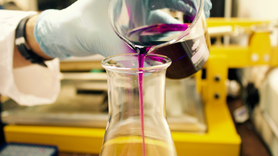 A person wearing a lab coat and blue gloves pours a purple liquid form one beaker into another