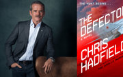 Man in formal clothing leaning on an arm chair and a book cover titled The Defector by Chris Hadfield.