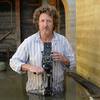 Gideon Mendel standing in water up to their waist, holding a retro camera.
