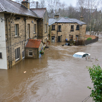 a residential area where flood water has risen; the flood water is almost reaching the downstairs floor windows and a white car is almost fully submerged.
