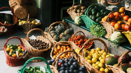 A variety of fruit and vegetables sit in several baskets outside a shop. These include avocado, mango, pineapple, apple, tomato, plum, pear and lemon baskets