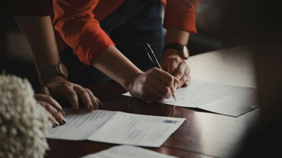 Two people stand over documents placed on a dark wood table. One writes on one piece of paper whilst the second person holds the other in place.