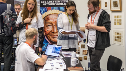 People stand and talk as one kneels to typr on a computer, surrounding a onfrence fair stand.