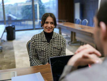Two people sitting at a desk facing eachother. One is looking at a laptop.