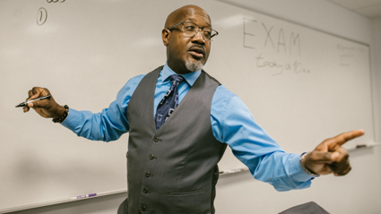 A teacher stands at the front of a classroom and speaks whilst writing on a whiteboard