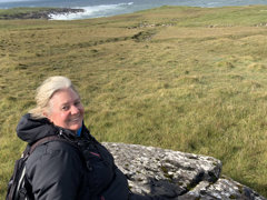 Jo Bollard sitting on a large rock in the middle of a grassy cliff overlooking the sea.