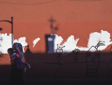 Woman carrying child in front of a terracotta wall. The wall features drawings which are implied to have been drawn by children.