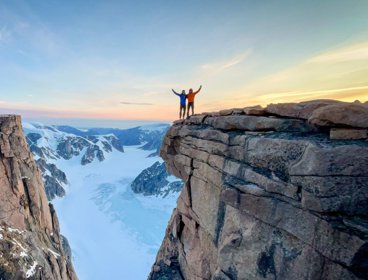 Two people in mountain gear stood at a snowy cliff edge at sunrise.