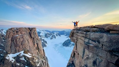 Two people in mountain gear stood at a snowy cliff edge at sunrise.