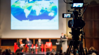 A panel of speakers on stage viewed from the back of a theatre, with a camera filming the event in the foreground.