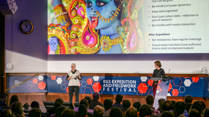 Two speakers on a stage at the RGS expedition and fieldwork festival. A slideshow with tips on how to be mindful during expeditions is projected on the wall behind them.