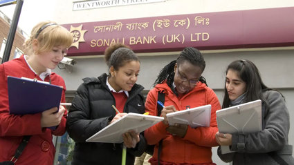 A group of three students with clipboards, collecting information in a high-street area. They are standing outside a bank.
