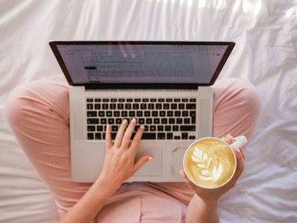 A Laptop on a persons lap with a coffee in their right hand