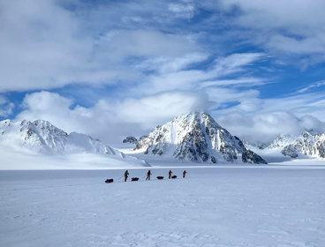 Four people trek across a snowy landscape with sleds, surrounded by towering snow-covered mountains.