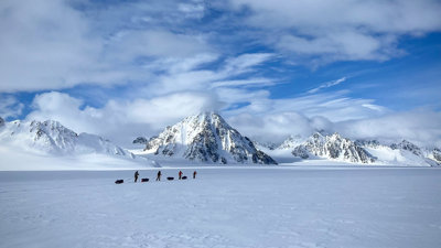 Four people trek across a snowy landscape with sleds, surrounded by towering snow-covered mountains.