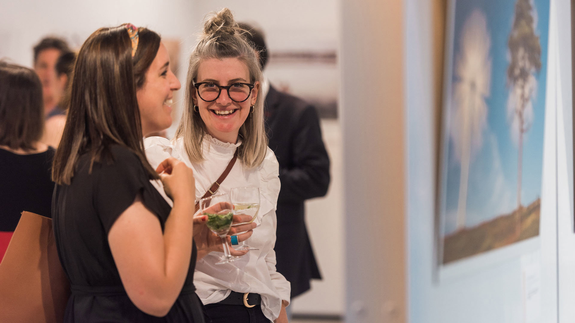 Two young people holding a drink while at a photograph exhibition. One person smiles directly at the camera.