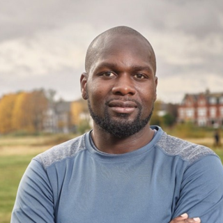 Headshot photograph of Dwayne Fields standing in a field with their arms crossed.