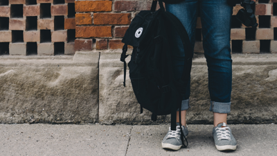 A High school student stands against a brick wall whilst holding their backpack in their hands by their side.