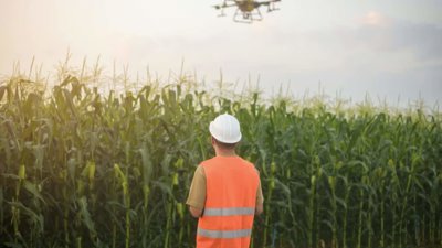 Person in high-vis vest controlling a drone in the sky over grassland .