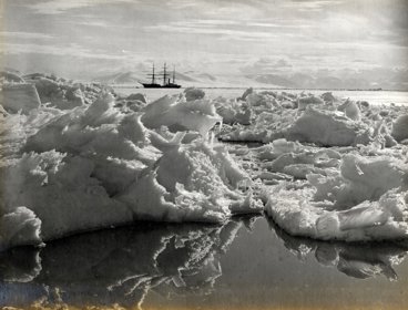Black and white photo taken in Antarctica of ice in the foreground and a large vessel on the water in the background