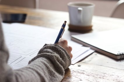 Person sat a table holding a pen and writing on a large piece of paper. On the table there is a mug on a coaster and a notebook.