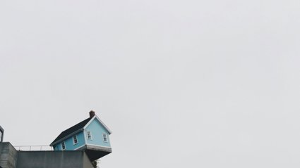 Small blue house precariously perched on the edge of a concrete building against a gray sky.