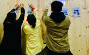 Three people seen from behind as they pin or clip printed pages and photos onto a string against a wooden wall. 