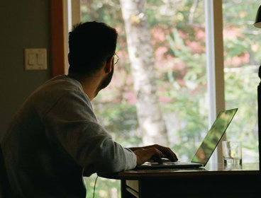 A person working on a laptop, facing away and staring out a window.