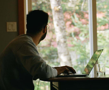 A person working on a laptop, facing away and staring out a window.