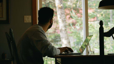 A person working on a laptop, facing away and staring out a window.