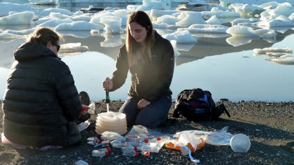 Two people sat beside a glacial lake taking measurements. They are surrounded by small transparent tubes and other measuring equipment.