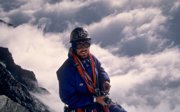An expeditioner in climbing gear on the summit of a mountain with a blanket of clouds below him.