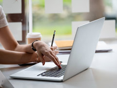 Person using a laptop at a desk with a notebook, pencil, and coffee cup nearby in a bright, modern setting.