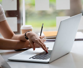 Person using a laptop at a desk with a notebook, pencil, and coffee cup nearby in a bright, modern setting.