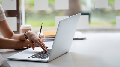 Person using a laptop at a desk with a notebook, pencil, and coffee cup nearby in a bright, modern setting.