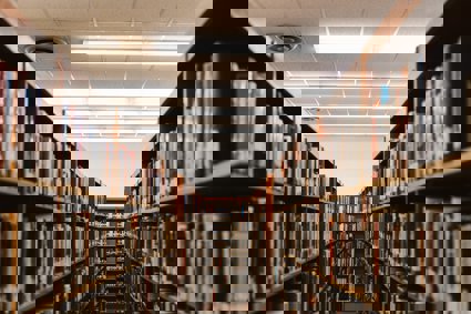 Two rows parallel libary shelves with a perpendicular row the can be seen between the two. All shelves are stacked full of books.