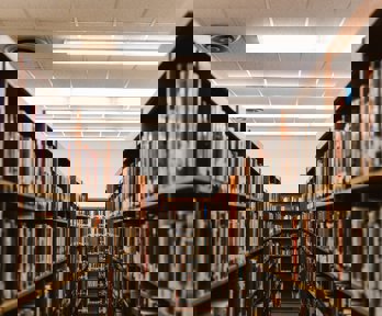 Two rows parallel libary shelves with a perpendicular row the can be seen between the two. All shelves are stacked full of books.