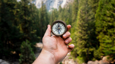 A hand holding a compass with a backdrop of mountains and trees.