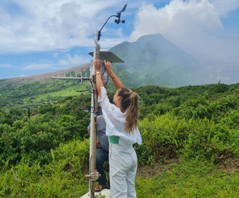 Two people with an air quality monitoring device in St George's Hill, Montserrat. Volcanic plume rises in the background.