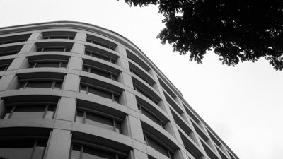A modern building seen from below. In the top corner are some tree branches. The photo is in black and white.