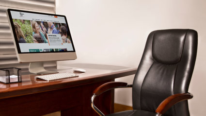 A leather office chair with wooden arm rests and a desk. A monitor on the desk is displaying the RGS website.
