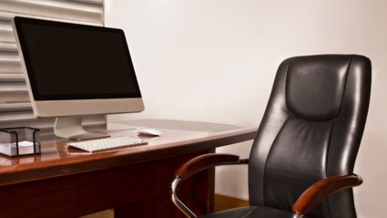 A leather office chair with varnished brown wood arm rests sit empty on front of a brown wooden desk. A laptop, keyboard, mouse  and a stack of note paper sit on the desk.
