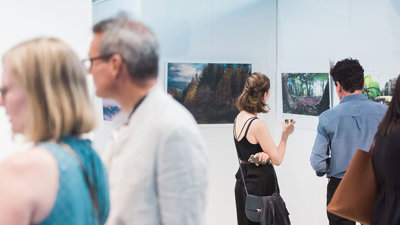 People viewing a photographic exhibition displayed on white panels in a modern pavilion. The photos show forests and trees.