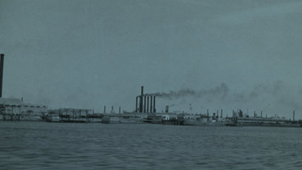 Black and white photograph of a river in the foreground and a factory in the background.