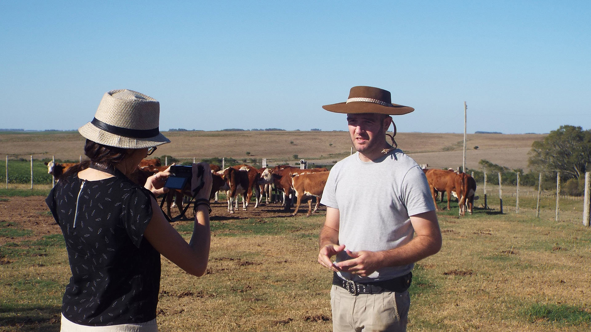 A person with a camera is filming another person wearing a cowboy hat. They are on a cattle farm in open farmland, with cows standing in the background.