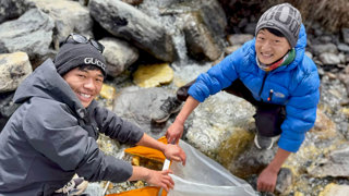 Two smiling people in outdoor gear use a net in a rocky mountain stream, collecting samples in cold weather.