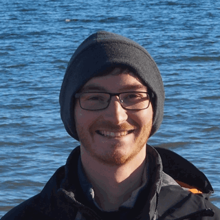 Headshot of Julian Martin. He is outside on a sunny day. Behind him is a a calm sea. 